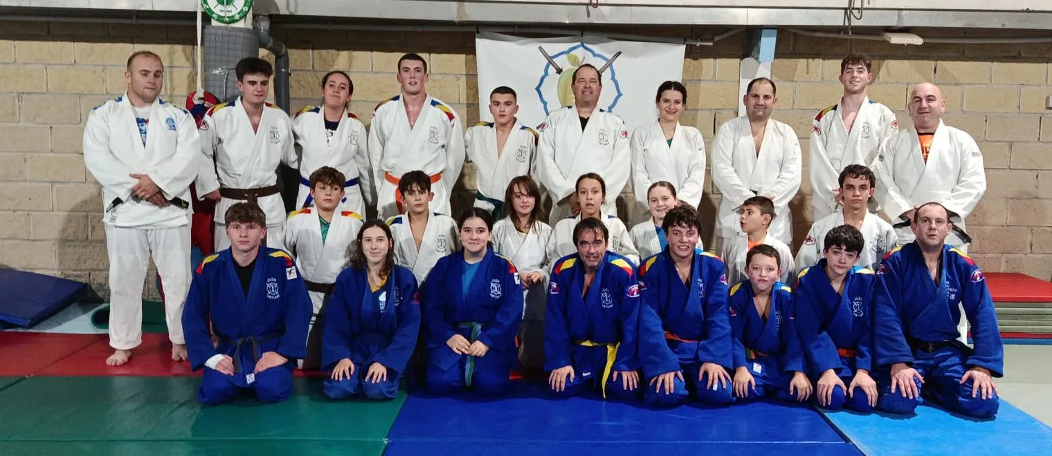 Foto de grupo de Judo Astures en el tatami: judokas de todas las edades, con judogi blanco y azul, junto a sus entrenadores y delante de la bandera del club